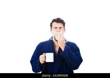 Portrait der verspäteten junge Mann trägt blau Bademantel holding Tasse Kaffee und Gähnen nicht in der Lage, rechtzeitig zur Arbeit zu kommen, auf einem weißen backgroun isoliert Stockfoto