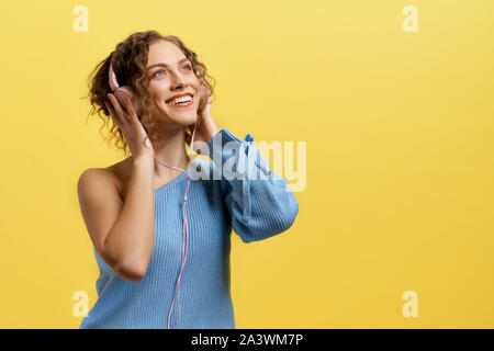 Ansicht von der Seite der bezaubernden Mädchen Tragen von Kopfhörern über und genießen Sie Musik auf gelb isoliert Hintergrund im Studio suchen. Aktive helle Frau hören Lieder und Lachen. Konzept der Unterhaltung. Stockfoto