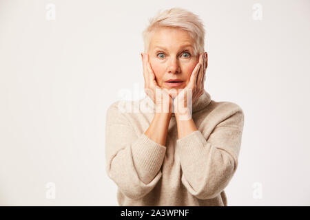 Portrait von schockiert reife Frau mit kurzen blonden Haaren halten Sie Ihr Gesicht mit den Händen, und wenn man die Kamera auf weißem Hintergrund Stockfoto