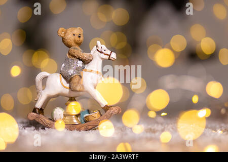 Dekorative Figuren aus einem Thema Weihnachten. Figur eines kleinen Teddybär auf einem Schaukelpferd. Weihnachtsdekoration. Festliches Dekor, warme Bokeh li Stockfoto