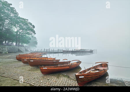 Dawn Cloud beginnt zu zerstreuen, da scheint die Sonne durch den Nebel über rudern Boote am Ufer am See Derwentwater, Lake District, Cumbria günstig Stockfoto