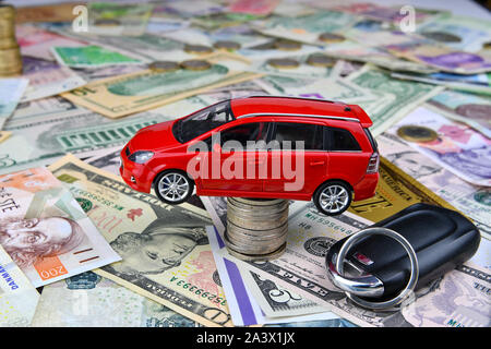 Ein autoschlüssel und ein rotes Spielzeugauto auf einen Turm aus Münzen. Hintergrund der verschiedenen nationalen Währung und ein symbolisches Gold Dollar Banknote. Konzept des c Stockfoto