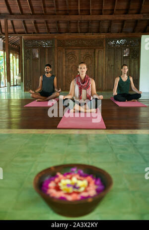 Mixed-Race Gruppe der eine Frau und zwei Männer sitzen auf Yogamatten innen traditionelle indonesische Tempel auf Bali Üben Yoga zusammen. Stockfoto