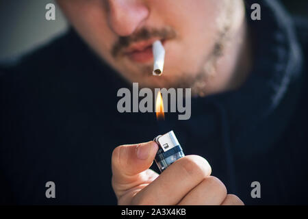 Ein bärtiger Mann in schwarzer Kleidung brennt eine Zigarette im Mund mit einem gasfeuerzeug. Stockfoto