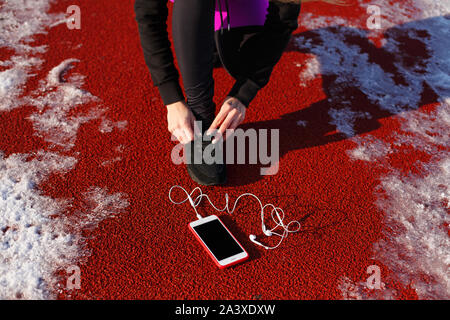 Mädchen Athlet in schwarzen Sneakers, geduckt auf der roten Spur ausgeführt werden. In der Nähe ist ein Telefon mit drahtgebundenen Kopfhörern. Kalten verschneiten Wetter. Stockfoto