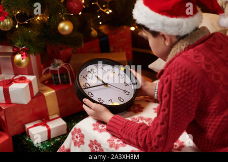 Teen Boy wartet auf Santa und beobachtete die Uhr, Hallenbad in der Nähe von eingerichteten Xmas Tree mit Lichtern, gekleidet wie santa Helper - Frohe Weihnachten und Ha Stockfoto