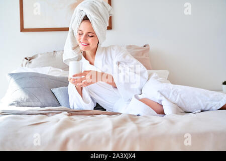 Foto junge Frau im Bademantel mit Tasse Kaffee in der Hand liegend auf Bett im Schlafzimmer Stockfoto