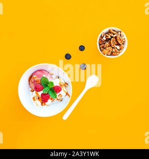 Vanille- und Erdbeereis mit Karamell, Himbeeren und Minze auf weiße Schüssel mit Muttern und Heidelbeeren auf orange gelb hinterlegt. Schöne desse Stockfoto