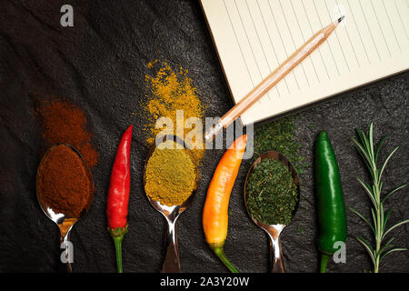 Verschiedene Gewürze auf einem schwarzen Stein und gelbe Notebook für Rezept mit Bleistift. Orientalische Gewürze in Löffel und Rosmarin, Pfeffer auf Schwarz metalli Stockfoto