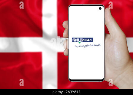 Ein Mann hält ein Telefon mit dem Logo der Firma Chr. Hansen Holding aufgeführt im OMX Kopenhagen. Dänische Flagge Hintergrund. Credit: PIXDUCE Stockfoto