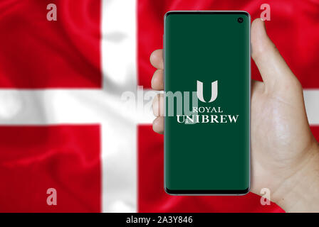 Ein Mann hält ein Telefon mit dem Logo des Unternehmens Royal UNIBREW aufgeführt im OMX Kopenhagen. Dänische Flagge Hintergrund. Credit: PIXDUCE Stockfoto