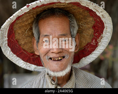 Älteren burmesischen Mann mit schlechten Zähnen trägt eine traditionelle asiatische konische Hut und Lächeln für die Kamera. Stockfoto