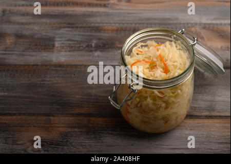 Hausgemachten Sauerkraut mit Karotten in einem Glas mit einem offenen Deckel auf einem Holz- braunen Hintergrund. Im rustikalen Stil Stockfoto