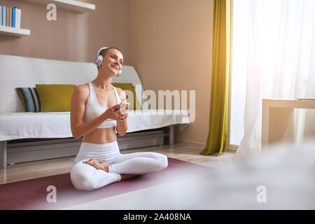 Kaukasischen jungen kurzhaarige Frau lächeln und sitzen auf Matten in Licht und Musik mit weißen Kopfhörer anhören. Genießen yoga Stockfoto