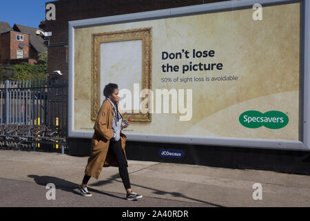 Eine Dame geht an einem grossen Plakat ad für High Street Augenoptiker Kette, Specsavers, am 2. Oktober 2019, in London, England. Stockfoto