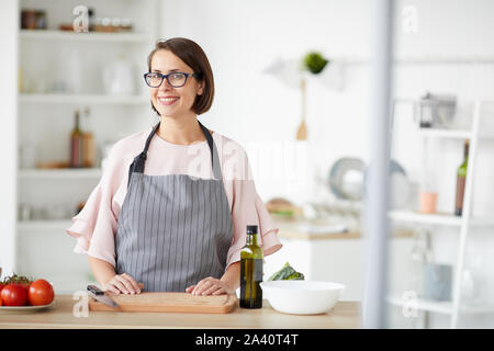 Porträt der jungen Frau in Brillen tragen Schürze steht in der Nähe der Küche Tisch und ein Lächeln auf die Kamera in der Küche Stockfoto