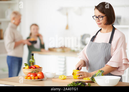 Junge Frau in Brillen tragen Schürze Schneiden von Gemüse auf Schneidebrett für Abendessen, während in der Küche stehen mit ihrer Familie im Hintergrund Stockfoto