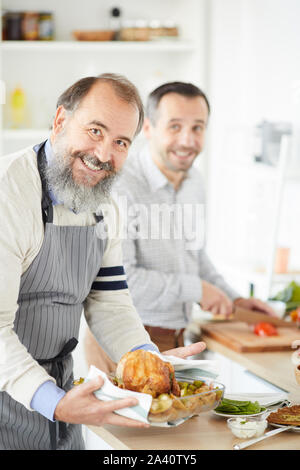 Portrait von älteren Mann mit Bart lächelt in die Kamera während der Vorbereitung Truthahn mit jungen Mann helfen ihm im Hintergrund Stockfoto
