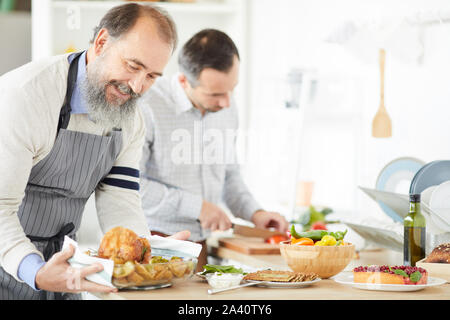 Älterer Mann mit Bart Vorbereitung Rost der Türkei für Abendessen, während Jungen Mann Schneiden von Gemüse für Salat im Hintergrund in der Küche Stockfoto