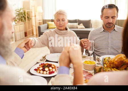 Schönes Paar mit geschlossenen Augen am Esstisch halten sich an den Händen und beten mit den anderen Mitgliedern der Familie zu Hause sitzen Stockfoto