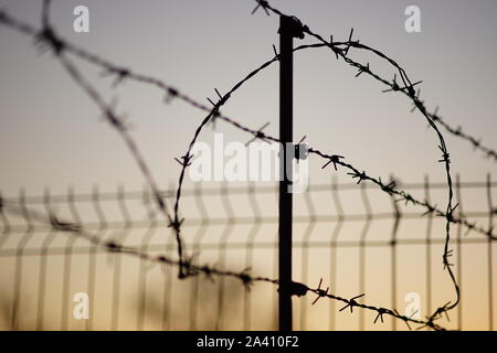 Schwarze Silhouette von einem Stacheldrahtzaun im Sommer abends, hellblau orange Himmel Hintergrund. Stockfoto
