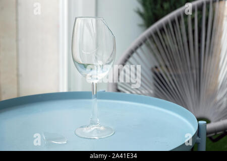 Gebrochene leeren Becher. Ein Glas Becher auf einem Bein steht auf einem kleinen Tisch auf der Terrasse. Ein Stück Glas liegt in der Nähe des gerissenen Glas Wein. Stockfoto