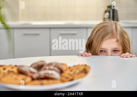 Selektiver Fokus der gerissen kleine Mädchen verstecken sich hinter Tisch in Küche und köstliche Schokolade Cookies bleiben auf dem Tisch. Kleines Kind wünschen süße Muffins. Konzept der Süßigkeiten und Essen. Stockfoto