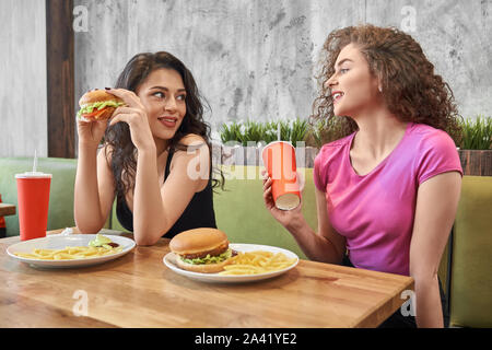 Ansicht von der Seite der zwei hübschen Mädchen essen leckere Hamburger und Trinken süßen Wasser im Cafe. Weibliche Freunde sich einander, reden und miteinander kommunizieren. Konzept der Mahlzeit und Fast Food. Stockfoto