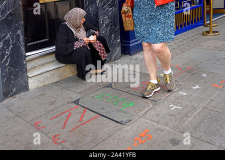 London, England, UK. Asiatische Frau mit Kopftuch in einem Türrahmen sitzend, eine Frau vorbei gehen. Stockfoto