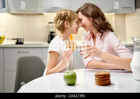 Seitenansicht hübsche Mutter und Tochter cookies essen und trinken Milch in der Küche. Frauen und Mädchen am Tisch sitzen, an einander suchen, Lachen und berühren mit Nasen. Konzept der Liebe.