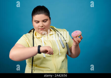 Pralle Brünette junge Mädchen halten Donut auf ihre Hand und sah auf ihre Uhr auf der anderen Seite. Fetten weiblichen Versucht nicht ungesund zu Essen und Sport mit einem Seil um den Hals hängend zu tun Stockfoto