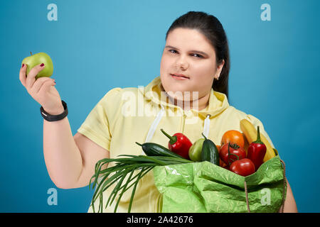 Seite der Brünette übergewichtige Mädchen halten große grüne Paket mit frischem Obst und Gemüse auf ihre Hand, die Kamera im Studio. Hübsche junge Frau in gelb Pullover kaufen Nahrung für gesunde Diät Stockfoto