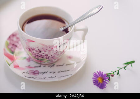 Schwarzer Tee mit Löffel in einem eleganten Oldtimer porzellan Schale auf dem weißen Tisch. Zarte Blume Chrysantheme in der Nähe. Stockfoto