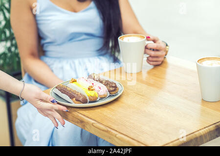 Weibliche Hand dient eine Platte der eclairs auf einem Tisch in einem Café. Das Mädchen am Tisch mit Eclairs, eine Tasse Kaffee in der Hand. Stockfoto