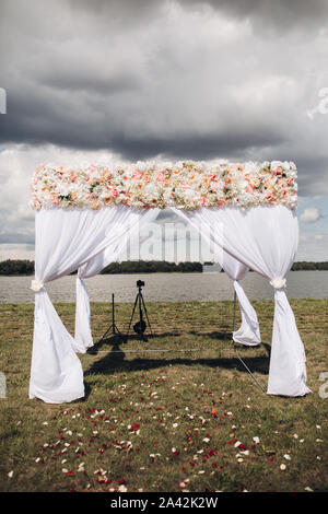 Blick über White Wedding arch mit Blumen auf der Oberseite und Rosenblätter auf dem Rasen verstreut. Durch den Fluss an bewölkten Tag gelegen. Stockfoto