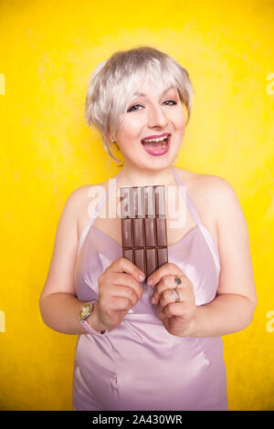 Süße blonde kurze Haare Frau mit dem Übergewicht in den Händen halten, eine große Tafel Schokolade auf Gelb, helles Studio Hintergrund Stockfoto