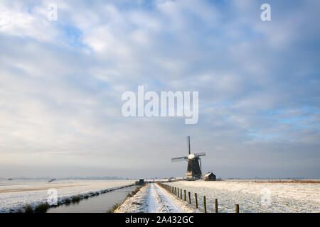 Blick auf die holländische Landschaft im Winter mit einem Pfad mit Zaun und einem Graben zu einer Windmühle, bedeckt mit Schnee unter netten Wolken in den Niederlanden. Stockfoto