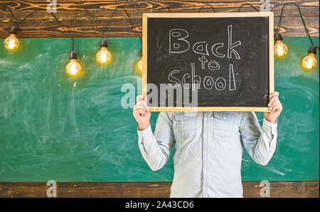 Beginn des Schuljahres Konzept. Lehrer hält Schiefertafel vor Gesicht. Mann begrüßt Studenten, schwarzes Brett für den Hintergrund. Lehrer gesichtslosen hält Tafel mit Titel zurück in die Schule. Stockfoto
