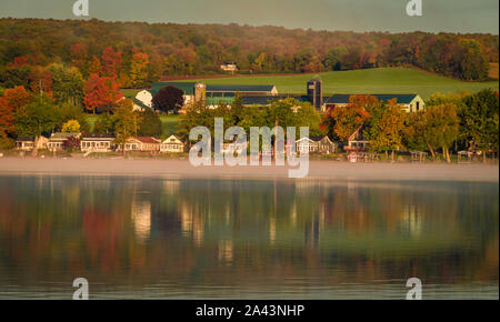 Nebel steigt über den See bei Sonnenaufgang mit Lakefront cottages Punktierung der Küstenlinie im Herbst Laub umgeben Stockfoto