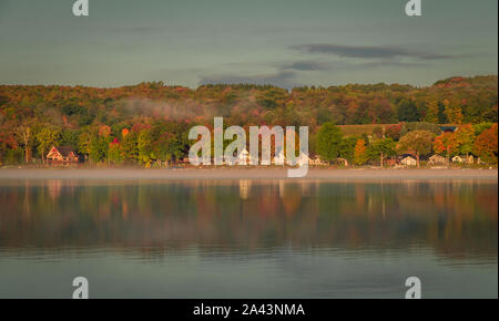 Nebel steigt über den See bei Sonnenaufgang mit Lakefront cottages Punktierung der Küstenlinie im Herbst Laub umgeben Stockfoto