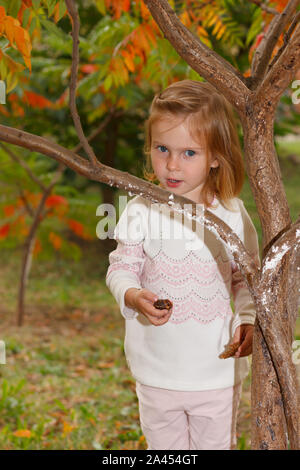 Adorable Baby Mädchen hält Schnecke, spielen in einem sonnigen Park unter einem Baum mit gelben Blätter, versteckt hinter Baum Stockfoto