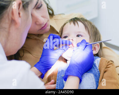 Der Arzt schaut auf die Zähne von einem kleinen Kind. das Baby in die Arme der Mutter sitzt. Zahnarzt untersuchen junge Zähne in der Klinik. Eine kleine Patienten in Stockfoto