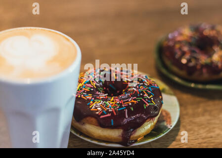 Schlechtes Frühstück. Kaffee mit einem gezeichneten Herz und Milch auf einen hölzernen Tisch in einem Coffee Shop. zwei Schokolade Donuts mit Streuung auf den Tisch neben der Co Stockfoto