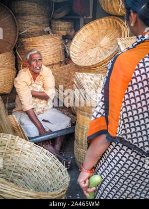 MYSURU (MYSORE), Karnataka/Indien - 15 Februar 2018: Am Devaraja Market ein geflochtener Korb Verkäufer spricht mit einer Frau als sitzt er durch seine Waren umgeben. Stockfoto