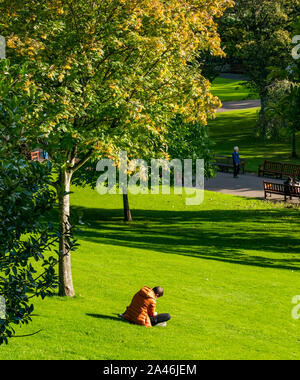 Die Princes Street, Edinburgh, Schottland, Großbritannien, 12. Oktober 2019. UK Wetter: Menschen in Edinburgh einen schönen warmen sonnigen Herbsttag in der Hauptstadt genießen. Ein Mann sitzt auf dem Gras lesen Stockfoto