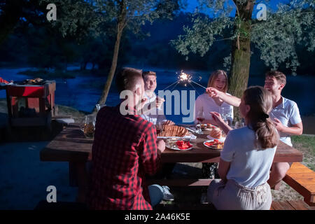 Gruppe von Freunden feiern gerne Urlaub mit Sprinklern und trinken Rotwein beim Picknick französischen Dinner Party im Freien in der Nähe des Ri Stockfoto