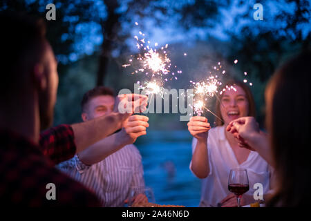 Gruppe von Freunden feiern gerne Urlaub mit Sprinklern und trinken Rotwein beim Picknick französischen Dinner Party im Freien in der Nähe des Ri Stockfoto