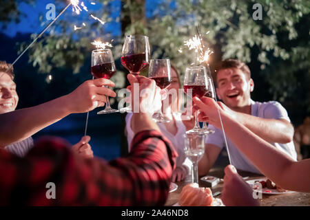 Gruppe von Freunden feiern gerne Urlaub mit Sprinklern und trinken Rotwein beim Picknick französischen Dinner Party im Freien in der Nähe des Ri Stockfoto