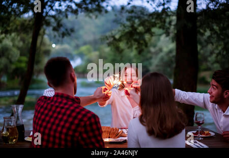 Gruppe von Freunden feiern gerne Urlaub mit Sprinklern und trinken Rotwein beim Picknick französischen Dinner Party im Freien in der Nähe des Ri Stockfoto