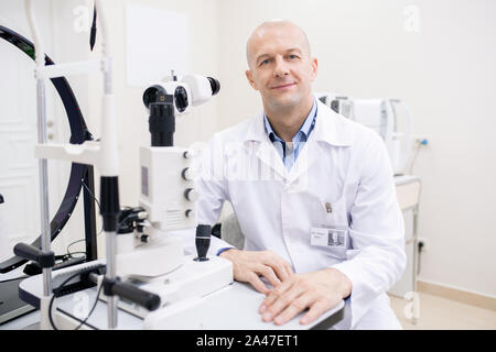 Gerne kahl Mitte - gealterte Augenarzt in Jungtieren durch Arbeitsplatz sitzen Stockfoto